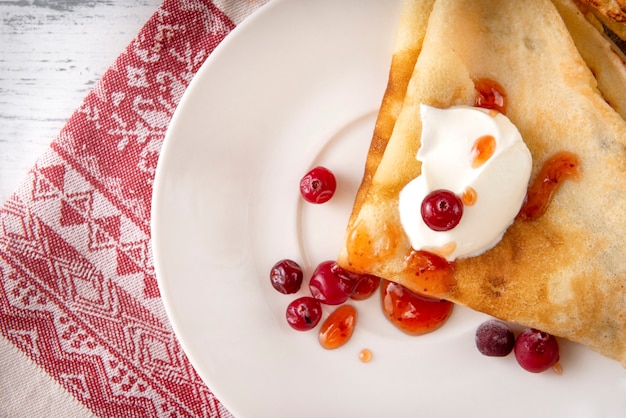 Russische Pfannkuchen mit saurer Sahne, Beeren, Preiselbeeren und Marmelade auf einem Teller, Tee, Marmelade auf einem Handtuch mit einem roten Muster auf einer weißen Oberfläche, Draufsicht,