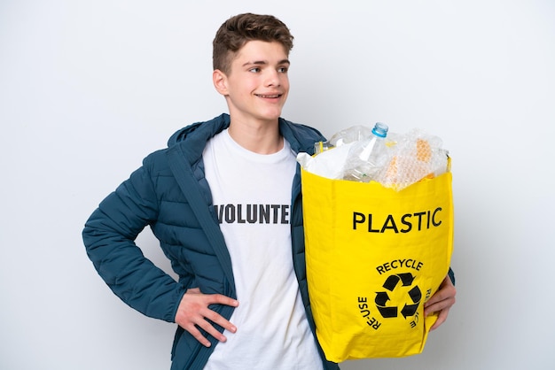 Russischer Teenager, der eine Tüte voller Plastikflaschen hält, um sie auf weißem Hintergrund zu recyceln, posiert mit Armen an der Hüfte und lächelt