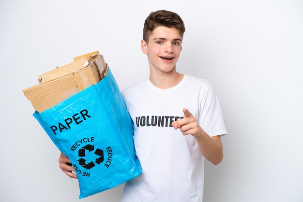 Russischer Teenager, der einen Recycling-Beutel voller Papier hält, um isoliert auf weißem Hintergrund zu recyceln, überrascht und zeigt nach vorne
