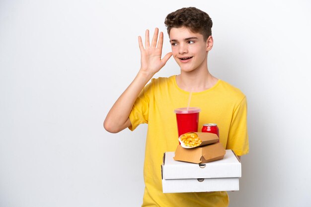 Russischer Teenager, der Fast Food isoliert auf weißem Hintergrund hält und etwas hört, indem er die Hand auf das Ohr legt
