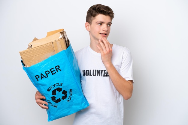 Russischer Teenager-Mann, der einen Recycling-Beutel voller Papier hält, um isoliert auf weißem Hintergrund zu recyceln und lächelnd nach oben zu schauen