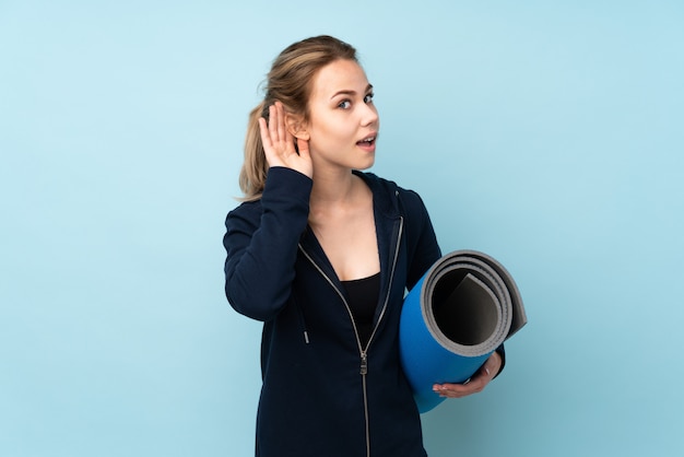 Russisches Mädchen des Teenagers, das Matte an der blauen Wand hält, die etwas hört, indem man Hand auf das Ohr legt