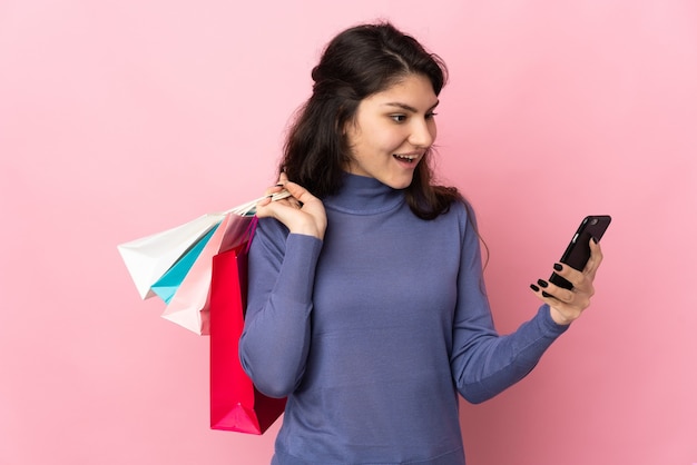 Russisches Mädchen des Teenagers lokalisiert auf rosa Wand, die Einkaufstaschen hält und eine Nachricht mit ihrem Handy an einen Freund schreibt