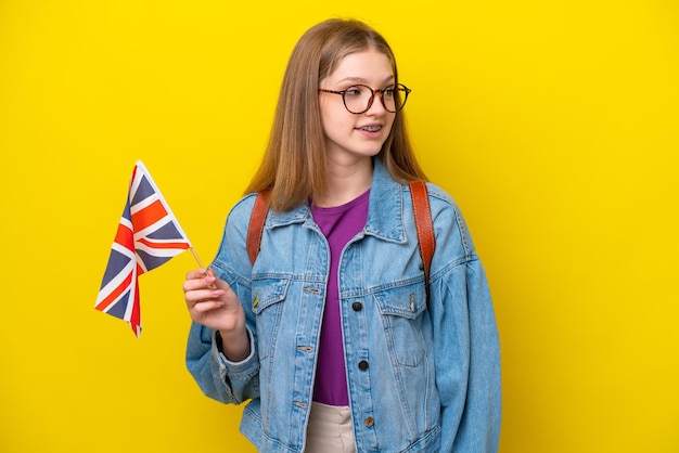 Russisches Mädchen im Teenageralter, das eine Flagge des Vereinigten Königreichs isoliert auf gelbem Hintergrund hält, zur Seite schaut und lächelt