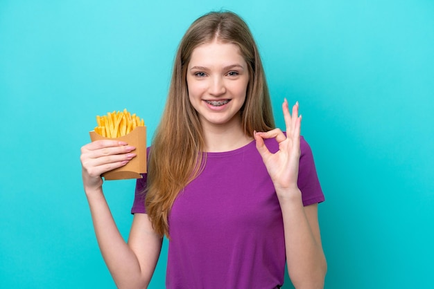 Russisches Mädchen im Teenageralter, das Pommes frites fängt, isoliert auf blauem Hintergrund, das mit den Fingern ein Ok-Zeichen zeigt