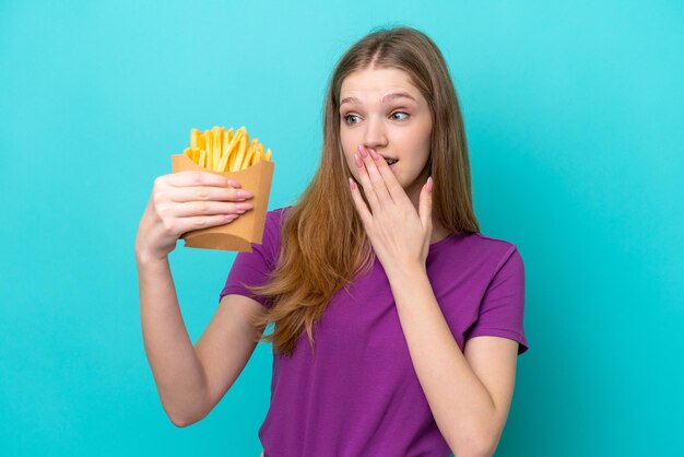 Russisches Mädchen im Teenageralter, das Pommes frites fängt, isoliert auf blauem Hintergrund mit Überraschung und schockiertem Gesichtsausdruck