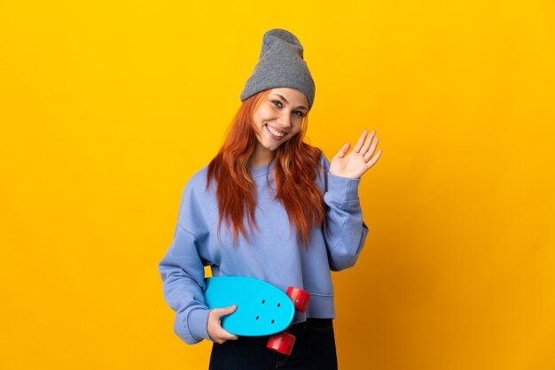 Russisches Teenager-Skatermädchen lokalisiert auf gelber Wand, die mit Hand mit glücklichem Ausdruck salutiert