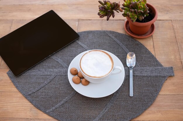 Una rústica mesa de madera lista para el desayuno con un capuchino caliente y unas galletitas. Tableta negra sobre la mesa