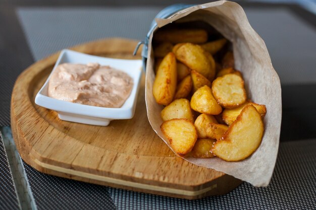 Rustikal gebratene Kartoffelspalten in einem Papierumschlag mit Sauce auf einer Holzplatte auf der Außenterrasse in einem Café.