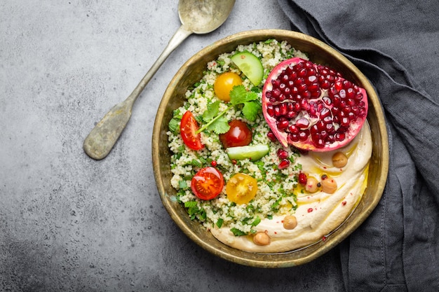 Rustikale Schüssel mit Couscous-Salat mit Gemüse, Hummus und frisch geschnittenem Granatapfel. Mahlzeit im nahöstlichen oder arabischen Stil mit Gewürzen und frischem Koriander. Gesundes mediterranes Abendessen, Platz für Text