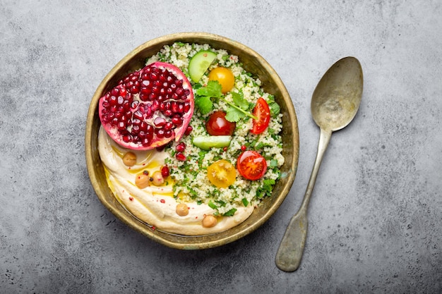 Rustikale Schüssel mit Couscous-Salat mit Gemüse, Hummus und frisch geschnittenem Granatapfel. Mahlzeit im nahöstlichen oder arabischen Stil mit Gewürzen und frischem Koriander. Schönes und gesundes mediterranes Abendessen
