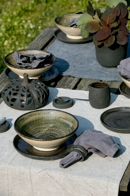 Rustikale Tischdekoration draußen im Garten mit leerem Keramikgeschirr, schwarzen Tellern und rauen Schalen, Kürbisdekorationen, auf Leinentischdecke über altem Holztisch. Gartenparty