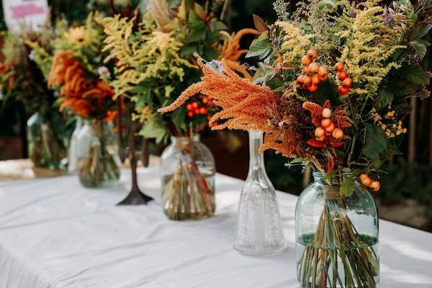 Rustikaler Stil. Blumensträuße im Boho-Stil auf dem Tisch in Glasvasen und Gläsern. Holztisch und weiße Tischdecke