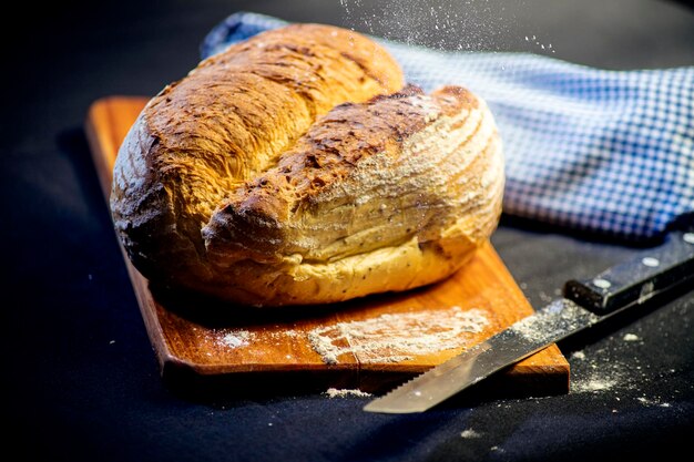 Rustikales Brot mit Mehl auf Holzbrett und Messer zum Schneiden von Brot auf schwarzem Hintergrund