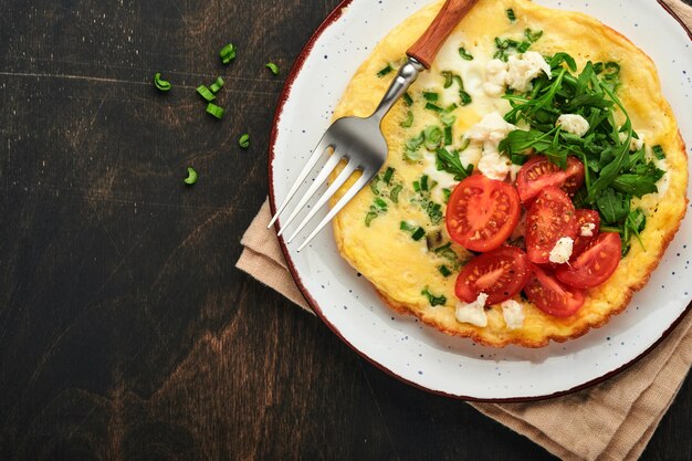 Rustikales Omelett oder Frittatas mit Frühlingszwiebeln, Käse, Mozzarella, grünem Rucola und Tomaten auf altem, dunklem Holzhintergrund. Gesundes Ernährungskonzept Frühstück Kopierbereich Draufsicht Mock up