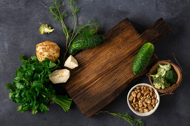 Rustikales Schneidebrett aus Holz mit Zutaten zum Kochen von veganem Essen auf Draufsicht des dunklen Hintergrunds