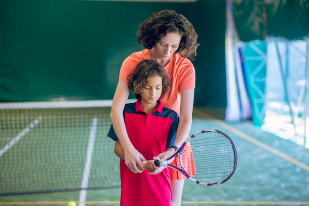 Rutina de ejercicio. Entrenador femenino en ropa brillante enseñando a un niño a sostener una raqueta de tenis