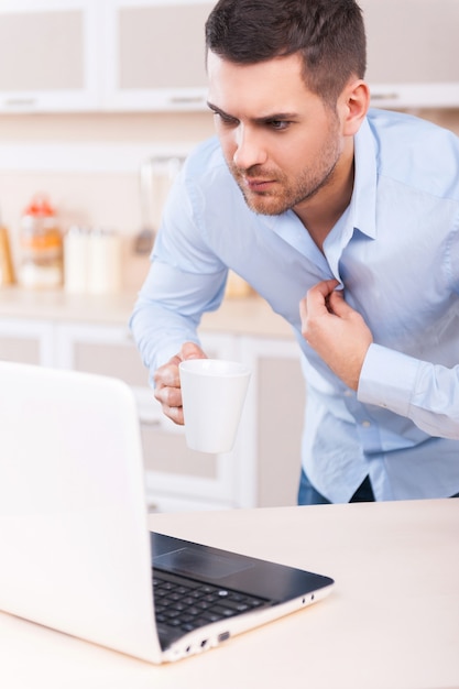 Rutina de la mañana. Hombre joven preocupado ajustando su camisa y mirando la computadora portátil mientras está de pie en la cocina