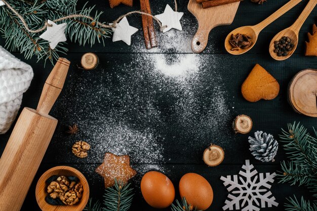 Árvore de Natal, biscoitos de gengibre, acessórios de panificação e ingredientes na mesa preta com farinha polvilhada. Copie o espaço