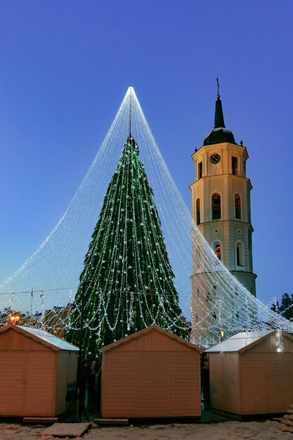 Árvore de Natal com decoração e barracas de Natal na Praça da Catedral em Vilnius, na Lituânia. Tem luzes de fada como se fosse um véu de noiva. Iluminado à noite