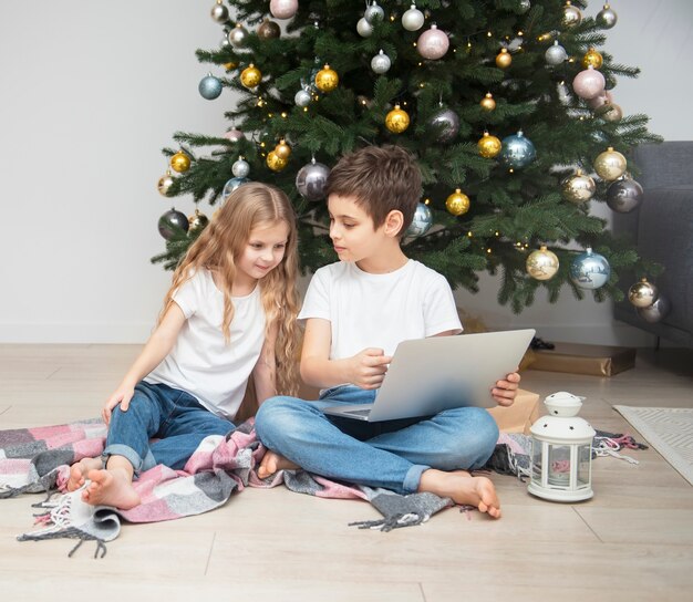 Árvore de Natal em uma grande sala de estar. As crianças brincam perto da árvore de Natal. Um menino com um laptop se comunica com os amigos.