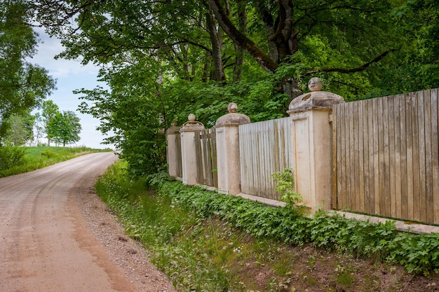 Árvore e cerca ao longo de uma estrada de terra perto de Nogale Manor Letónia Cerca de madeira com colunas de pedra e chão