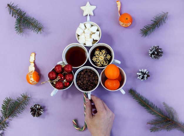 Árvore festiva feita de xícaras de chá cheias de chá, café, grãos de café, açúcar, bolas de Natal, tangerinas, galhos de pinheiro, em um fundo pastel na mão, vista de cima, bokeh