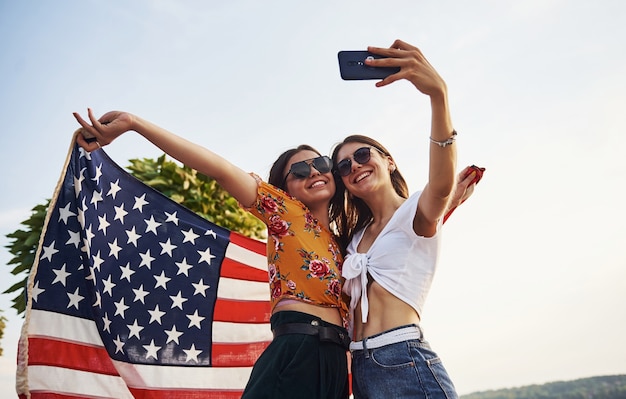Árvore verde no fundo. Duas mulheres alegres patrióticas com a bandeira dos EUA nas mãos, fazendo selfie ao ar livre no parque.