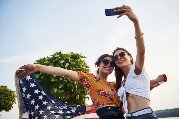 Árvore verde no fundo. Duas mulheres alegres patrióticas com a bandeira dos EUA nas mãos, fazendo selfie ao ar livre no parque.