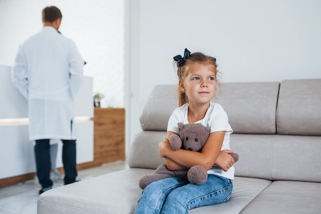 Ärzte dahinter. Nettes kleines Mädchen mit Teddybär in den Händen sitzt im Wartezimmer des Krankenhauses.