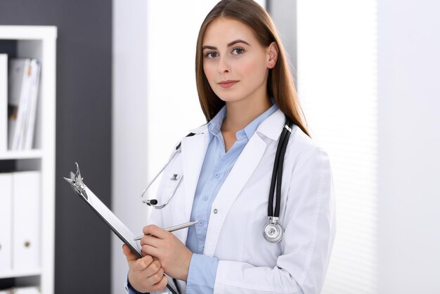 Ärztin füllt medizinisches Formular aus, während sie in der Nähe des Fensters im Krankenhausbüro steht. Glücklicher Arzt bei der Arbeit. Medizin- und Gesundheitskonzept.