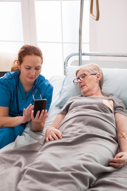 Ärztin hilft alter Frau, die mit dem Handy auf dem Bett sitzt. Alte Frau im Pflegeheim.