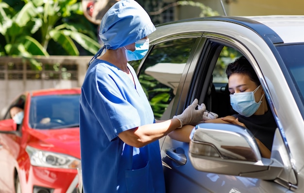 Ärztin in blauer Krankenhausuniform und Gesichtsmaske steht in der Nähe des Autos, das eine Spritze in den Händen hält, trägt Gummihandschuhe, die den Covid-19-Impfstoff injizieren, um in der Impfwarteschlange durch die Schulter des Patienten zu fahren.