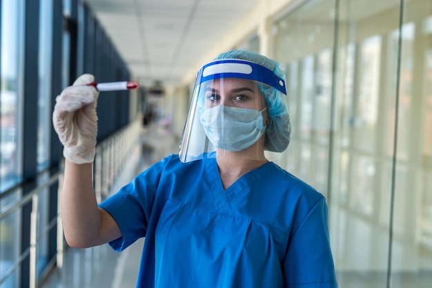 Ärztin in blauer Uniform Gesichtsmaske Gesichtsschutz und weißen Handschuhen halten Reagenzglas mit positivem Ergebnis für Covid 19. Pandemie-Konzept