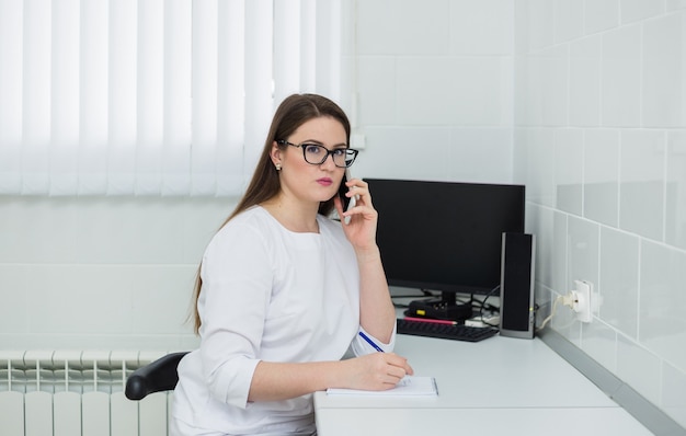 Ärztin mit Brille und weißem Mantel sitzt an ihrem Schreibtisch und telefoniert