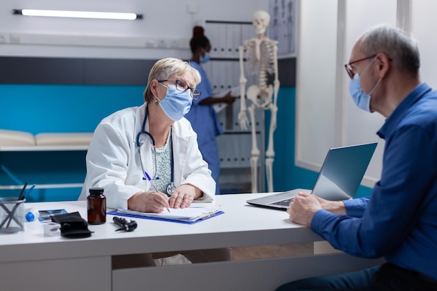 Ärztin mit Gesichtsmaske, die bei ärztlicher Beratung mit dem Mann Notizen zu Papieren macht. Arzt unterschreibt Zwischenablagedateien für medizinische Behandlung und Medizin während der Covid-19-Pandemie