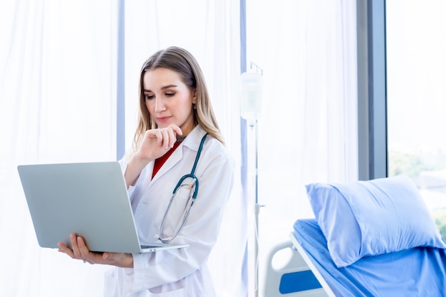 Ärztin mit Stethoskop, die mit Laptop arbeitet und auf Papierkram auf Holztisch im Krankenhaushintergrund schreibt