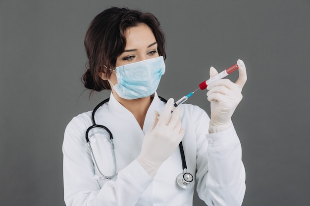 Ärztin mit Stethoskop in Maske, die Injektion hält, Gegenmittel gegen Coronavirus auf grauem Hintergrund.