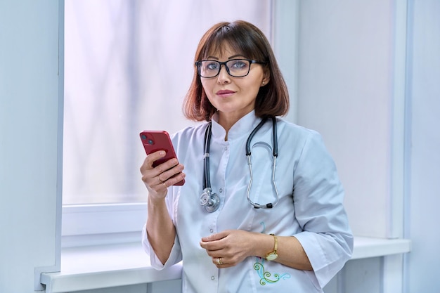 Ärztin mit Stethoskop und Smartphone in der Hand, die in ein Krankenhaus in die Kamera schaut.