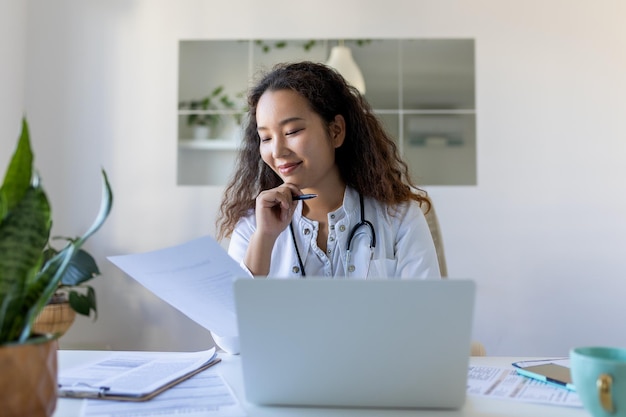 Ärztin, professionelle asiatische Ärztin, die Uniform trägt und sich Notizen in medizinischen Zeitschriften macht, die Dokumente über die Krankheitsgeschichte des Patienten füllen und auf den Laptop-Bildschirm schauen, Student, der sich ein Webinar ansieht