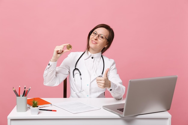 Ärztin sitzt am Schreibtisch und arbeitet am Computer mit medizinischer Dokumenthaltermünze im Krankenhaus einzeln auf pastellrosa Wandhintergrund. Frau im medizinischen Kittelbrillenstethoskop. Gesundheitsmedizinisches Konzept