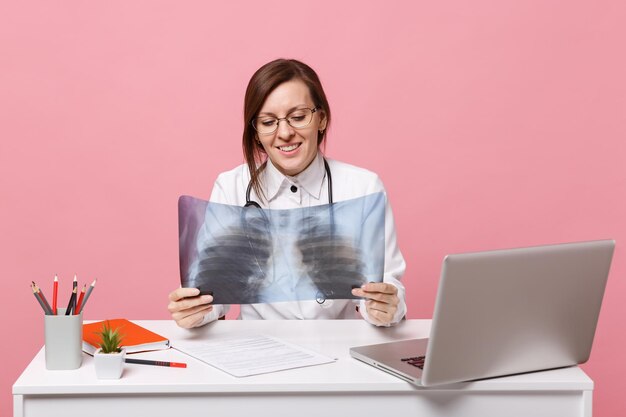 Ärztin sitzt an der Schreibtischarbeit am Computer mit medizinischem Dokument, das Röntgen im Krankenhaus isoliert auf pastellrosa Wandhintergrund hält. Frau im medizinischen Kittelbrillenstethoskop. Konzept der Gesundheitsmedizin.