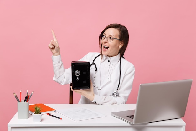 Ärztin sitzt an der Schreibtischarbeit am Computer mit medizinischem Dokument halten Banksafe im Krankenhaus einzeln auf pastellrosa Hintergrund. Frau im medizinischen Kittelbrillenstethoskop. Konzept der Gesundheitsmedizin.