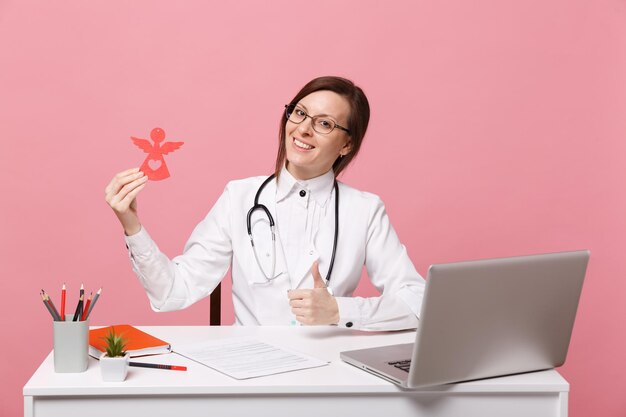 Ärztin sitzt an der Schreibtischarbeit am Computer mit medizinischem Dokument halten Engel im Krankenhaus isoliert auf pastellrosa Wandhintergrund. Frau im medizinischen Kittelbrillenstethoskop. Konzept der Gesundheitsmedizin.