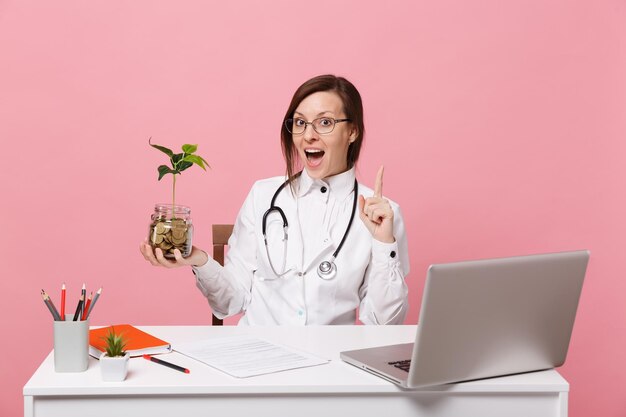 Ärztin sitzt an der Schreibtischarbeit am Computer mit medizinischen Dokumenten halten Münzen im Krankenhaus einzeln auf pastellrosa Wandhintergrund. Frau im medizinischen Kittelbrillenstethoskop. Konzept der Gesundheitsmedizin.