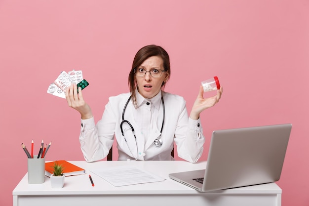 Ärztin sitzt an der Schreibtischarbeit am Computer mit medizinischen Dokumenten halten Pillen im Krankenhaus einzeln auf pastellrosa Wandhintergrund. Frau im medizinischen Kittelbrillenstethoskop. Konzept der Gesundheitsmedizin.