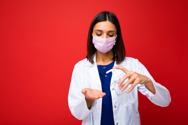 Ärztliche Beratung und Gesundheitsschutz während der Coronavirus-Epidemie. Junge Ärztin in Schutzmaske, weißer Kittel zeigt Antiseptikum in ihren Händen, lokalisiert auf Hintergrund, freier Raum, Studioaufnahme.