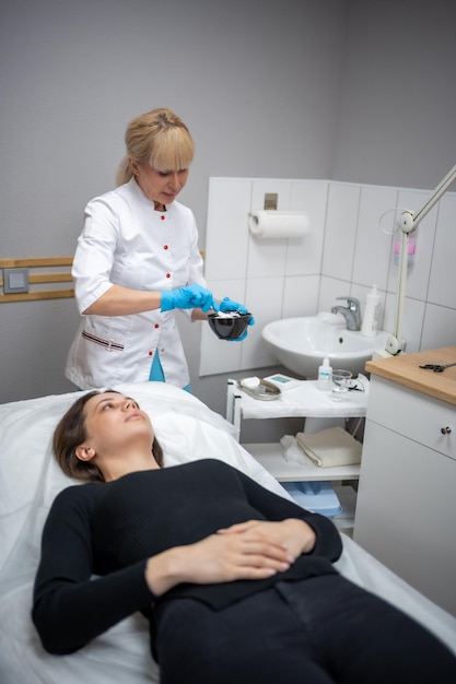 Ärztliche Kosmetikerin oder Dermatologin, die Gesichtsmaske im Kosmetiksalon herstellt Professionelle Kosmetikerin, die eine Gesichtsmaske auf das Gesicht einer kaukasischen Frau aufträgt, die im Bademantel auf dem Bett liegt