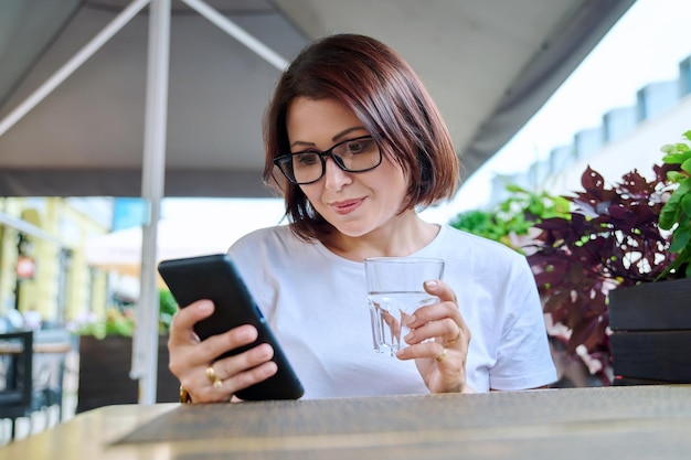 S-Frau sitzt in einem Café im Freien mit einem Glas Wasser mit dem Smartphone