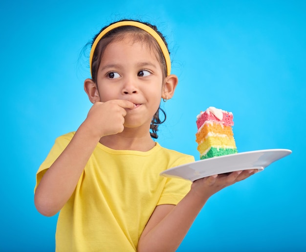 Sabor a pastel de cumpleaños y una niña con un dulce en el estudio comiendo glaseado para el postre del evento de la fiesta Comida arcoíris de celebración y niño hambriento en un estudio aislado y de fondo azul listo para celebrar